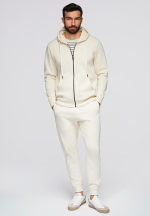 Mand med skæg iført cremefarvet hoodie, stribet skjorte, matchende joggingbukser og hvide sneakers, stående med hænderne i lommerne.