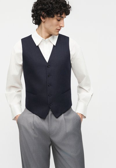 Άντρας με σγουρά μαλλιά, φορώντας λευκό πουκάμισο με κουμπιά, μπλε σκούρο γιλέκο και γκρι πλισέ παντελόνι, με τα χέρια στις τσέπες, κοιτάζοντας προς τα κάτω.