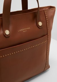 Brązowa skórzana torba tote z złotymi studami, przednia kieszeń oraz logo "Liebeskind Berlin" w złotych literach.