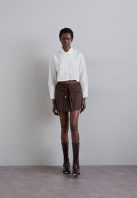 Chemisier blanc à manches courtes avec un devant plissé, associé à une mini-jupe marron avec un cordon rouge. Modèle portant des bottes marron jusqu'aux genoux.