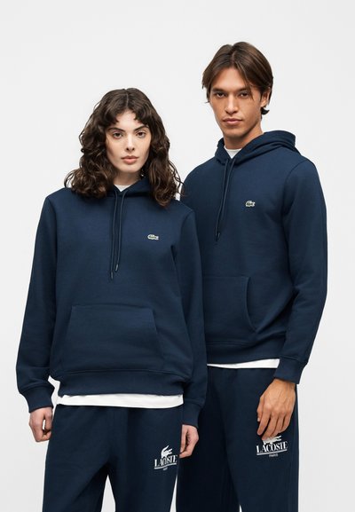 Два модела, облечени в еднакви тъмносини худита и спортни панталони Lacoste, стоят един до друг на едноцветен фон.