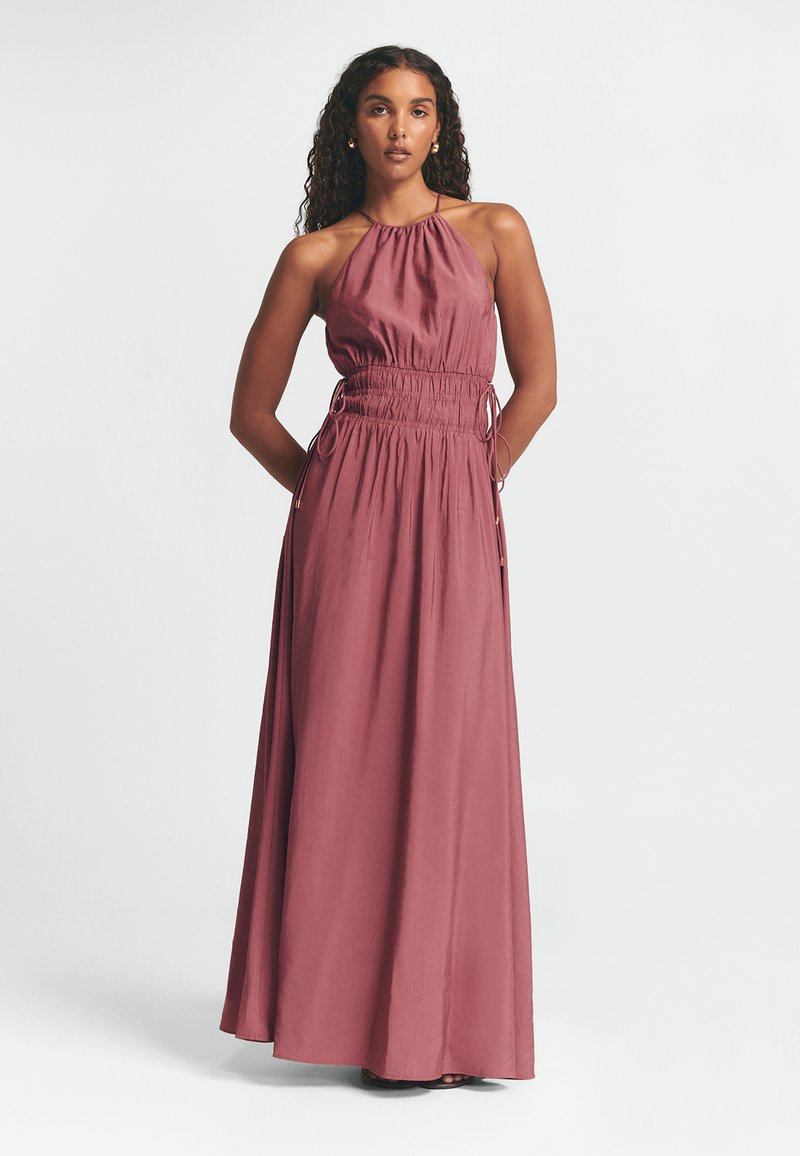 Maxi suknelė iš prislopintos rožinės, turinti kaklo raištelį, elastinę taliją ir šoninius raiščius su metaliniais akcentais. Minkšta, plaukianti medžiaga.