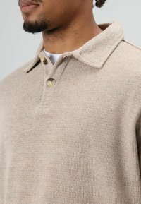 Polo en maille marron clair avec un col structuré, doté de deux boutons beiges et d'une texture douce. Gros plan sur l'encolure.