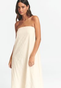Trägerloses, langes Kleid in Creme mit vertikaler Rippstruktur. Der weiche Stoff fällt elegant und präsentiert eine fließende Silhouette sowie ein minimalistisches Design.