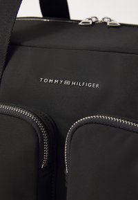 Sac en nylon noir avec fermetures éclair argentées et un logo, comprenant deux poches avant et une bandoulière détachable. Texture lisse et design minimaliste.