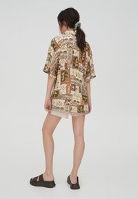 Femme avec queue de cheval portant une chemise ample à motifs, un short beige et des sandales noires, debout contre un fond uni, vue de dos.