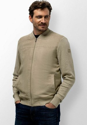 Man met kort bruin haar die een beige jas met rits en donkere spijkerbroek draagt, staand met handen in de zakken, kijkt iets naar de zijkant.