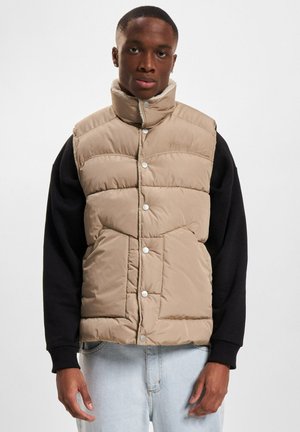 Beige Steppweste mit hohem Kragen, Druckknöpfen und gestepptem Design. Trägt man über einem schwarzen Langarmshirt, vor einem neutralen Hintergrund gezeigt.