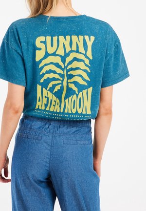 T-shirt en coton bleu-vert avec un effet dégradé, présentant un grand texte jaune et un motif de palmier au dos. Coupe décontractée, manches courtes.