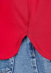 Rotes Baumwoll-T-Shirt mit abgerundetem Saum, ausgestattet mit seitlichen Schlitzen, kombiniert mit ausgebleichten Denim-Jeans, die eine kontrastierende Textur zeigen.