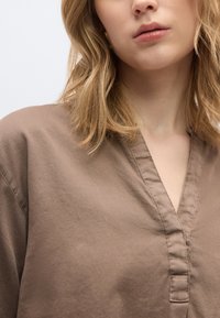 Ξανθός με καφέ μπλουζάκι με V λαιμόκοψη, με κυματιστά μαλλιά μήκους μέχρι τους ώμους, εμφανιζόμενος από τον λαιμό μέχρι το άνω μέρος του στήθους.