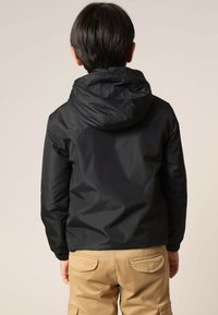 Veste imperméable noire avec capuche, poignets élastiques et texture lisse. Vue de dos, associée à un pantalon cargo de couleur claire.