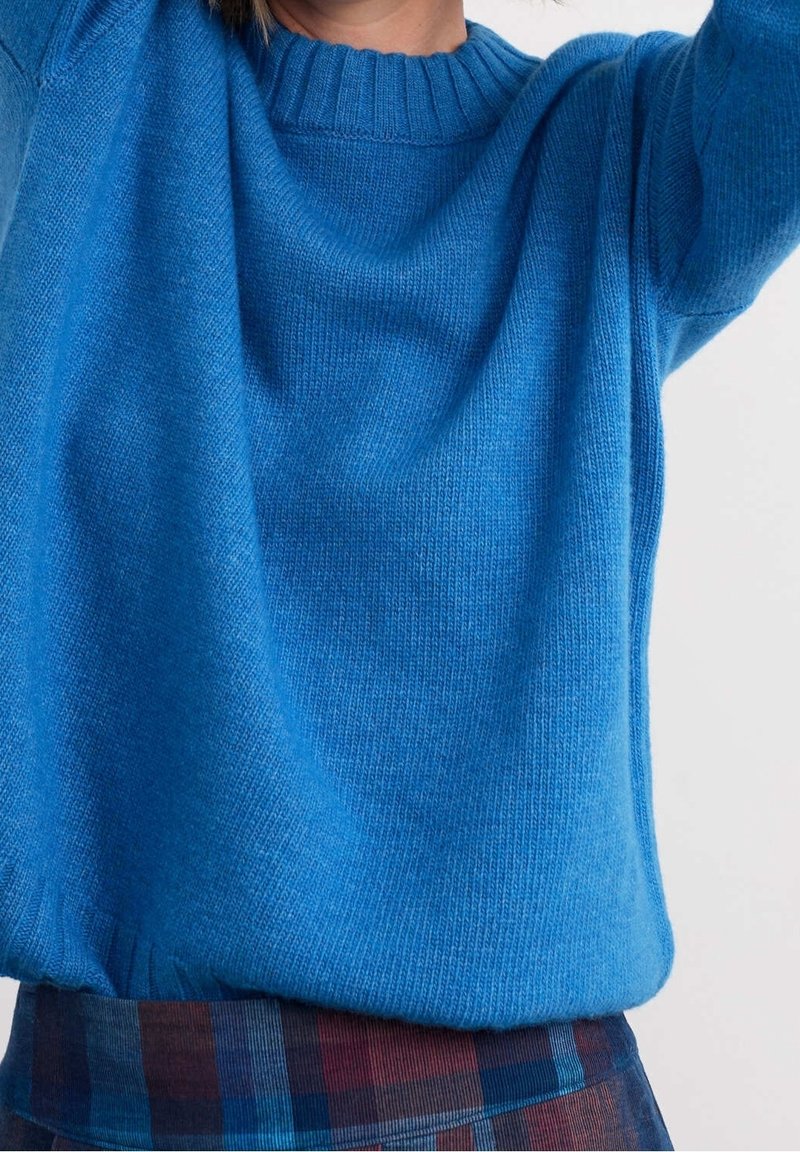 Personne portant un pull tricoté bleu ample et une jupe à carreaux avec les mains levées, cadrée au niveau du cou et de la taille sur un fond uni.