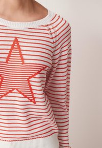 Pull en tricot avec des rayures rouges et blanches, présentant un grand motif d'étoile rouge sur le devant. Poignets et encolure côtelés.