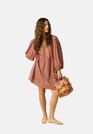 Femme aux longs cheveux ondulés portant une robe rose ample avec des motifs, tenant un sac à franges multicolore et portant des ballerines tissées beiges.