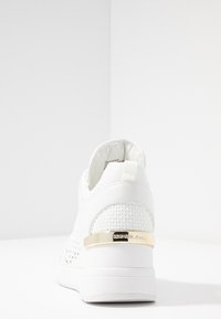 Sneakers bianchi con texture intrecciata, una piastra d'accento dorata sul tallone e un design elegante. La suola è di un bianco solido, offrendo un aspetto moderno.
