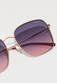 Ochelari de soare pătrați cu lentile gradient violet care trec în roz, cadru din metal auriu și brațe burgundii. Numele brandului "Vogue" este vizibil.