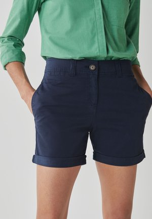 Personne portant un short bleu marine à ourlet roulé avec bouton et fermeture éclair à l'avant, les mains dans les poches, associé à une chemise verte à manches longues.