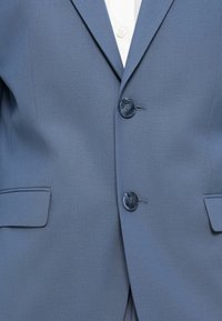 Μπλε σακάκι με υφασμάτινη υφή, δύο κουμπιά και πλαϊνές τσέπες, με λευκό πουκάμισο σε αντίθεση να φαίνεται από κάτω.