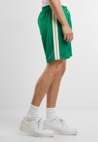 Grønne atletiske shorts med hvide sidelister, lavet af letvægtsmateriale. Har elastisk talje og lommer. Parret med hvide sneakers.