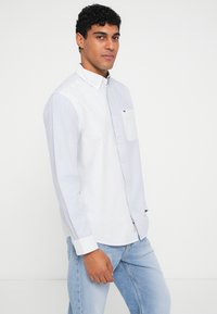 Γαλάζιο πουκάμισο με κάθετες ρίγες και σε μία πλευρά, μονόχρωμο από την άλλη, με γιακά που κλείνει με κουμπιά και τσέπη στο στήθος.