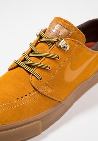 Baskets en daim orange Nike Stefan Janoski avec lacets rayés jaunes et noirs, semelle en caoutchouc marron et étiquette de languette brandée.