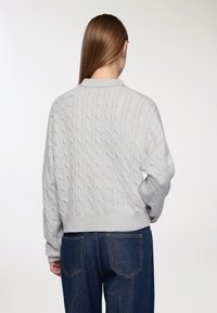 Kobieta z długimi brązowymi włosami nosi jasnoszary sweter w warkocz i ciemnoniebieskie dżinsy, zwrócona plecami na jednolitym białym tle.