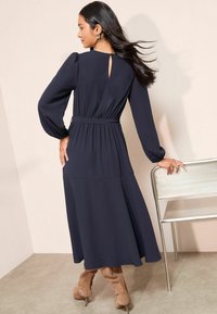 Jūras spēku zila garām piedurknēm kleita ar atslēgas caurumu aizmugurē, jostota vidukļa daļa un plūstoša svārku daļa; kombinēta ar pelēkām augstpapēžu kurpēm.