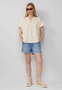 Cremfarbener, strukturiert kurzer Ärmeltop mit eingeschnittenem Kragen, kombiniert mit Jeansshorts und weißen Riemchensandalen, vor einem hellen Hintergrund stehend.