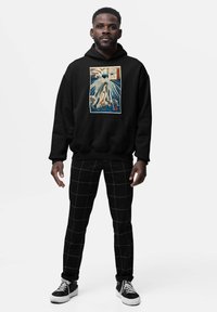 Man in een zwarte hoodie met een kunstprint van blauwe en beige golven, zwarte broek met een wit ruitpatroon en zwarte sneakers met witte zolen.