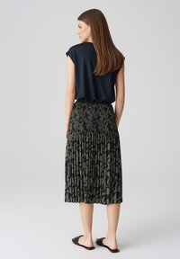 Top corto nero a maniche corte abbinato a una gonna midi plissettata verde con un motivo scuro. La modella indossa sandali piatti neri.