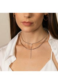 Doppeltgängiges Halsband mit quadratischen Metallperlen, ausgestattet mit einem vertikalen Kristallanhänger. Getragen über einem weißen Hemd mit Kragen.