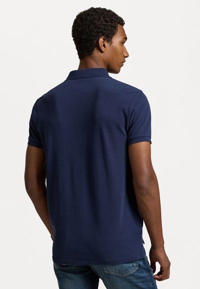 Polo de color azul marino con mangas cortas, cuello acanalado y aberturas laterales, hecho de una tela texturizada. Llevado con jeans azules.