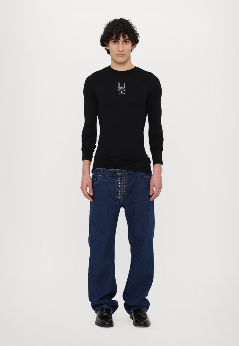 Schwarzes Langarmshirt mit einem Logo auf der Brust, kombiniert mit lockeren blauen Jeans, die eine einzigartige Schnürung an der Vorderseite haben, und schwarzen Schuhen.