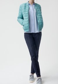 Veste bomber matelassée bleu clair avec fermeture éclair, portée par-dessus une chemise bleu clair à rayures verticales et un jean en denim foncé, assortie de baskets grises.