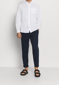 Camicia bianca in lino con bottoni e taschino, abbinata a pantaloni a gamba affusolata blu scuro e sandali neri con cinturini e fibbie.