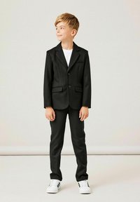 Czarny garnitur z marynarką jednorzędową, klapami w szpic, kieszeniami bocznymi i slim fit spodniami, zestawiony z białym T-shirtem i trampkami.