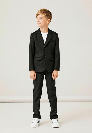 Costume noir avec veste à un bouton, revers en pointe, poches latérales et pantalon slim, associé à un t-shirt blanc et des baskets.