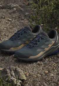 Green and gray athletic shoes with a textured upper, featuring blue accents and a beige stripe, designed for outdoor use.