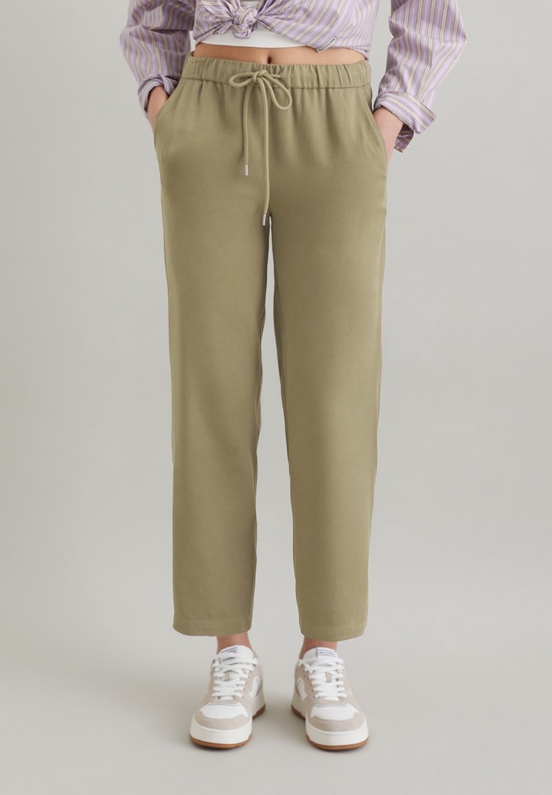 Olivgröna joggarbyxor med elastisk midja, snörning, sidofickor och en avslappnad passform, i kombination med vita sneakers.
