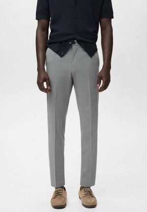 Uomo che indossa pantaloni eleganti aderenti grigio chiaro, scarpe in suede color nocciola con lacci e una camicia nera a maniche corte con i bottoni inferiori slacciati.