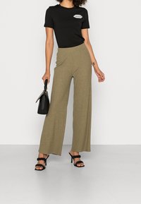 T-shirt en coton noir avec un logo, associé à un pantacourt taille haute à côtes vert olive et à des talons noirs à lanières. Tenant un sac à main noir.