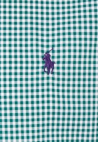 Grön- och vit rutigt tyg med en lila broderad polo ryttare logga i centrum. Texturerat bomullsmaterial.