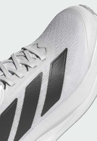 Weißer Sportschuh mit strukturiertem Obermaterial, ausgestattet mit drei schwarzen Streifen und passendem Schnürsenkel. Die Sohle ist glatt und weist eine leichte Kurve auf.