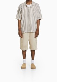 Randig kortärmad skjorta i beige och vitt, beigea cargoshorts, vita strumpor och bruna träskor. Enkel, avslappnad passform och funktionella fickor.