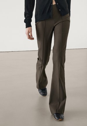 Personne portant un pantalon évasé à taille haute brun foncé et des chaussures pointues noires, marchant sur un sol lisse de couleur claire.