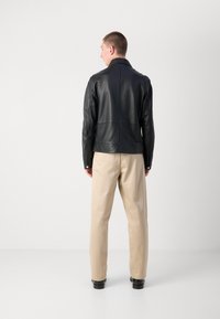 Jachetă din piele neagră cu un design minimalist, având un guler în picioare și o siluetă elegantă, purtată cu pantaloni bej și pantofi negri.