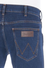 Niebieskie dżinsy z charakterystyczną tylną kieszenią ozdobioną beżową skórzaną naszywką, podkreśloną pomarańczowymi szwami i klasycznym zygzakowatym wzorem.
