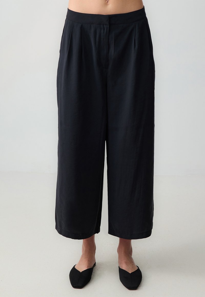 Pantalon noir large et court porté par une personne debout, associé à des chaussures noires sans lacets sur un sol clair.
