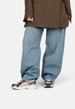 Persoon die lichtblauwe wijdbenige spijkerbroek, bruine en witte sneakers en een bruin colbert draagt, staand tegen een effen achtergrond.